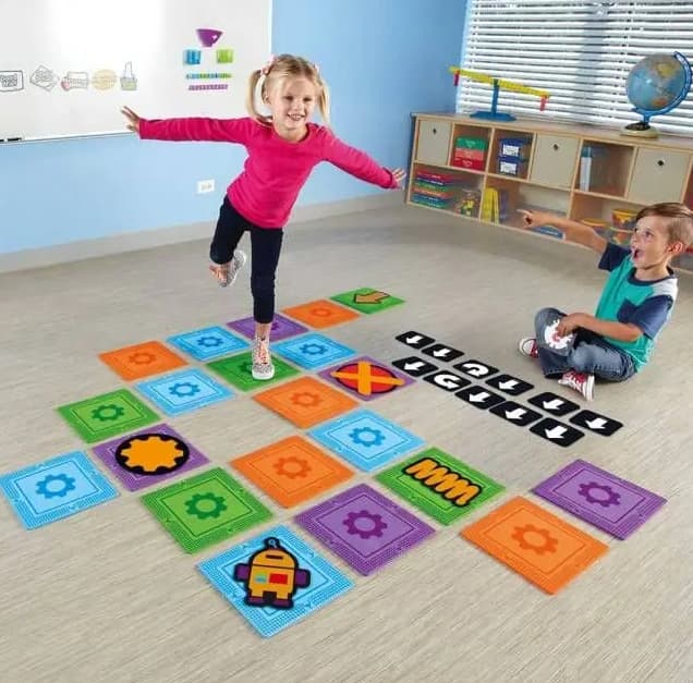 programmiamoci: bambini che giocano sul tappeto del coding. Bambina sopra al tracciato. Bambino a destra seduto